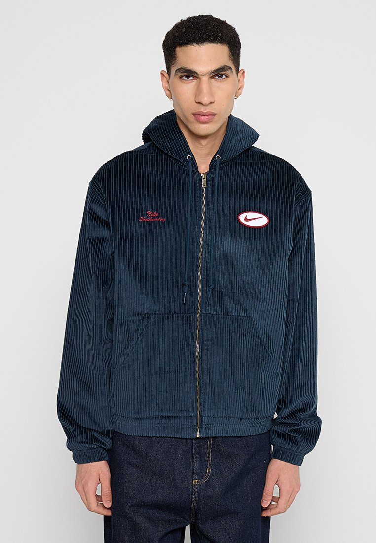 Haut à capuche en corduroy bleu marine avec fermeture éclair, poches avant, logo brodé en rouge et finition texturée. Comprend une capuche avec cordon de serrage.
