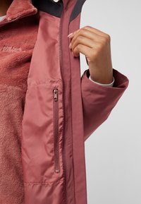 Veste pour femmes avec un intérieur en polaire maroon doux et texturé, un matériau extérieur lisse, une poche zippée et des manches ajustées.