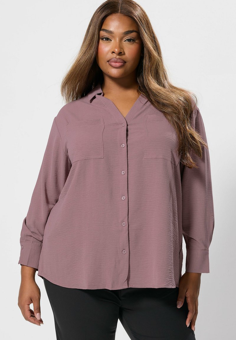 Camicia button-up di un tenue colore mauve con una vestibilità rilassata, maniche lunghe, due tasche sul petto e un tessuto testurizzato. Abbinata a pantaloni neri.