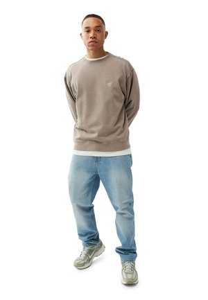Man standing with hands behind back, wearing beige sweatshirt, light blue jeans, and white and green sneakers on a white background.