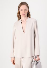 Blusa beige chiaro con maniche lunghe, scollo a V e design minimale. Il tessuto appare liscio, abbinata a una borsa grigia satinata.