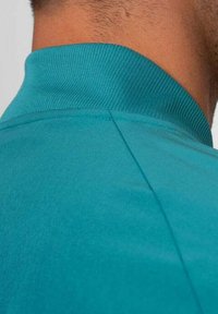 Camiseta polo color verde azulado con un cuello de canalé, que presenta una textura de tela suave y un detalle de costura a lo largo del hombro y la costura trasera.