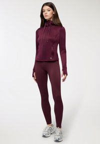 Giacca a zip piena color bordeaux con una texture liscia e un design aderente, abbinata a leggings coordinati e sneaker sportive argentate.