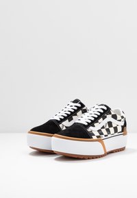Paire de baskets à plateforme Vans à damier noir et blanc avec des lacets blancs, des semelles épaisses blanches et des semelles extérieures en gomme brune sur un fond uni.