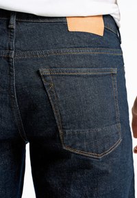 Dunkelblaue Denim-Jeans mit schlanker Passform, ausgestattet mit orangefarbenen Nähten, einer Gesäßtasche und einem beige Lederetikett, das mit "JUNK de LUXE" eingeprägt ist.