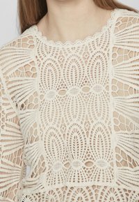 Top in pizzo con intricati motivi geometrici color crema, colletto ondulato e sezioni intagliate per un aspetto testurizzato e stratificato.