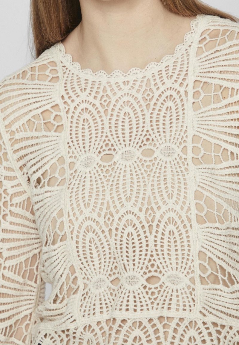 Top in pizzo con intricati motivi geometrici color crema, colletto ondulato e sezioni intagliate per un aspetto testurizzato e stratificato.