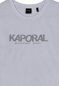 Camiseta de algodón blanca con un estampado de glitter plateado que dice "KAPORAL" y "sol de verano". Cuello redondo, mangas cortas, talla S.