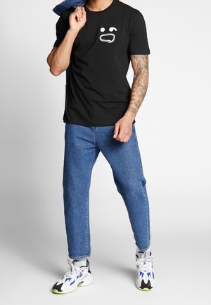 Man wearing black t-shirt with white carabiner design, blue jeans, white sneakers, holding blue jacket over shoulder against plain background.