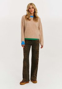 Kobieta z długimi blond włosami nosi beżowy sweter z niebieskimi i zielonymi wykończeniami, spodnie w panterkę oraz brązowe buty na platformie, stojąc na białym tle.