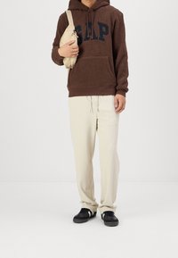 Hnědá fleecová mikina s kapucí a logem "GAP", krémové kalhoty a černé tenisky s bílými detaily. Drží malou béžovou kabelku přes rameno.