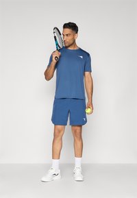 Blaues Sportshirt und Shorts aus leichtem Material, hält einen Tennisschläger und einen Ball. Weiße Sportschuhe und Socken vervollständigen das Outfit.