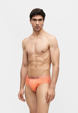 Muž s vypracovanou postavou stojaci na bielom pozadí, oblečený v jasne oranžových Emporio Armani slipkách, s vážnym výrazom, ktorý pozerá priamo pred seba.