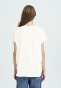 Kvinde med langt brunt hår iført en løs hvid kortærmet top og blå jeans, stående med ryggen til mod en ensfarvet baggrund.