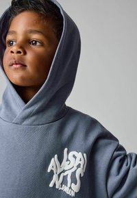 Bambino con pelle marrone e capelli scuri, che indossa un hoodie blu con il logo bianco "AllSaints London", che guarda a sinistra su uno sfondo grigio.