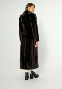 Dlouhý černý kabát z umělé kožešiny s límcem, s rovnými liniemi, měkkou texturou a bočními kapsami. Nosený přes robustní boty na podpatku.