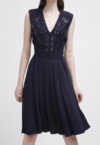 Marinblå klänning med djup V-ringning och ärmlös design, prydd med blommig broderi och paljetter på överdelen, utsvängd kjol, mjukt tyg.