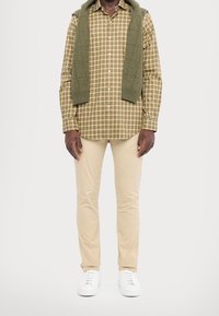 Man med beige byxor, vita sneakers, grön och brun rutad skjorta samt en olivgrön tröja som hänger över axlarna mot vit bakgrund.