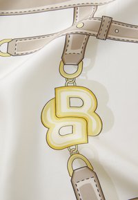 Stofă de mătase cu un design cu modele, având curele bej și un emblemă decorativă mare, aurie și albă, în formă de litere interconectate.