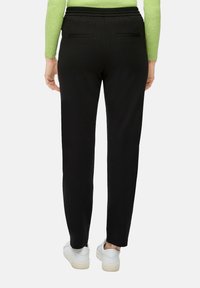 Pantaloni felpati neri a gamba affusolata con una vita elasticizzata, due tasche posteriori e una superficie liscia, abbinati a scarpe da ginnastica bianche.