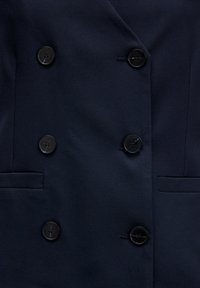Close-up van de donkerblauwe dubbele blazer met zes zwarte knopen en twee horizontale zakken.