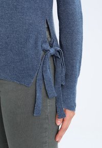 Maglione blu a coste con fessure laterali e accenti annodati. I dettagli includono maniche lunghe e tessuto testurizzato, abbinato a pantaloni verde oliva.