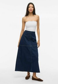 Gonna lunga in denim blu scuro con vita alta, caratterizzata da cuciture visibili e una forma svasata. Indossata con una maglietta bianca aderente e ballerine marroni.