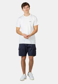 Camiseta de algodón blanca con un pequeño logo, combinada con pantalones cortos cargo azul marino con bolsillos laterales, y zapatillas blancas. Diseño simple y funcional.