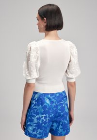 Maglietta bianca lavorata a maglia con maniche a sbuffo floreali texturizzate, abbinata a pantaloni corti blu con stampa floreale e orli arrotolati.