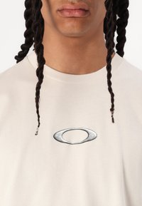 Beige cotton t-shirt with a rounded crew neckline, featuring a silver oval logo at center. Smooth texture and relaxed fit.
