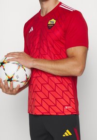 Homme portant un maillot de football rouge de l'AS Roma tenant un ballon de football Adidas blanc avec des motifs d'étoiles sur un fond uni.