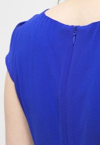 Blå klänning i mjukt, texturerat tyg. Har en rundad halsringning och en diskret dragkedjeslutning på ryggen. Korta ärmar.