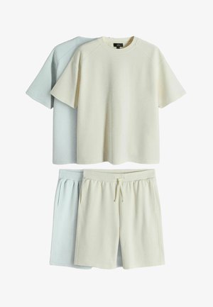 Zwei Sets kurzärmeliger T-Shirts und Shorts mit Kordelzug, eines in Creme und eines in Hellgrau, flach auf weißem Hintergrund ausgelegt.