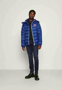 Casaco puffer azul com capuz, apresentando um fecho éclair branco, um logo cosido no peito e punhos canelados. Combinado com jeans escuros e botas pretas.