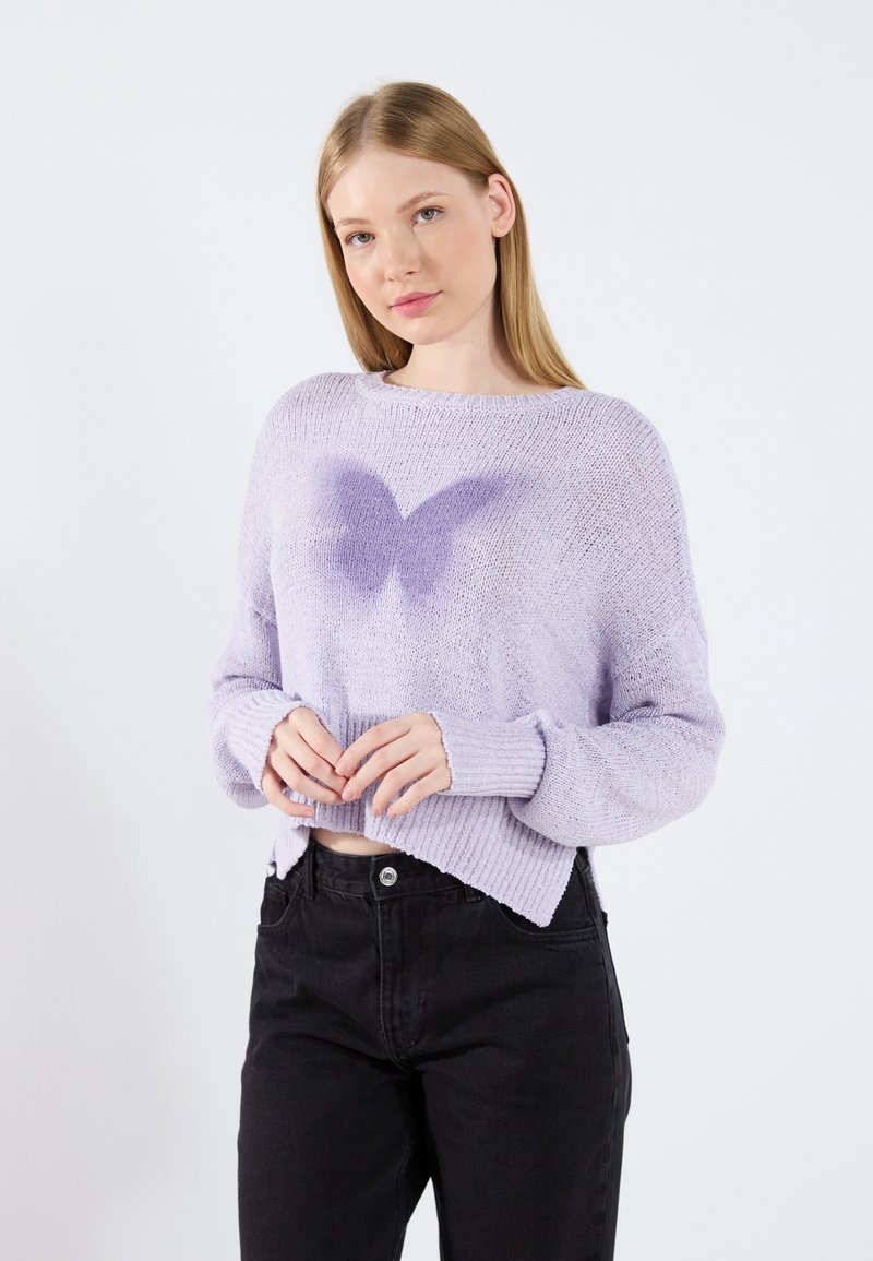 Pulover tricotat lila cu o croială lejeră și manșete ribbate. Are un design cu fluturi în lila mai închis la mijloc. Este asortat cu blugi negri cu talie înaltă.