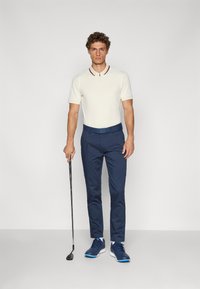 Golf obleka, ki vključuje kremasto polo majico z rdečim ovratnikom, mornarsko modre hlače in modre športne čevlje. Moški drži golf palico.