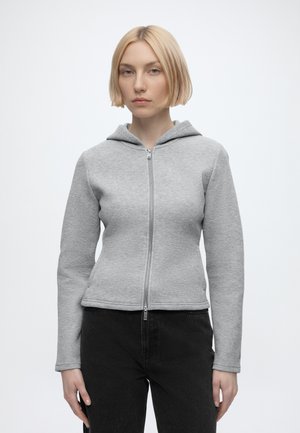 Donna bionda che indossa una felpa con cappuccio grigio chiaro con zip e pantaloni neri, in piedi davanti a uno sfondo bianco semplice.