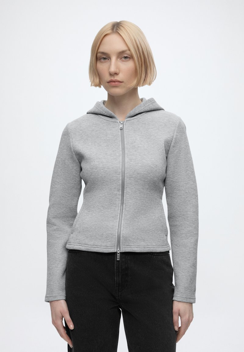 Femme blonde portant un sweat à capuche zippé gris clair et un pantalon noir, debout devant un fond blanc uni.