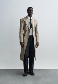 Beiger zweireihiger Mantel mit Spitzrevers über einem weißen Hemd, schwarzer Krawatte und dunklen Jeans aus Denim. Schwarze Schuhe mit glattem Finish.