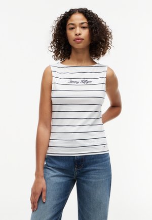 Femme aux cheveux bouclés portant un haut blanc sans manches à rayures bleu marine avec le texte "Tommy Hilfiger", associé à un jean bleu.