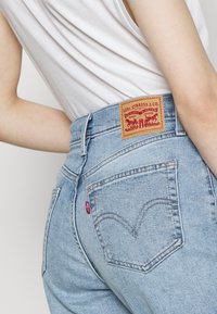 Ljusblå högmidjade jeans i denim, med en läderlapp med varumärke, röd etikett och klassisk sömnad på bakfickorna.