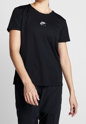 Kobieta ubrana w czarną siateczkową koszulkę Nike Air z krótkim rękawem i czarne spodnie, stojąca na tle gładkiego jasnego tła.
