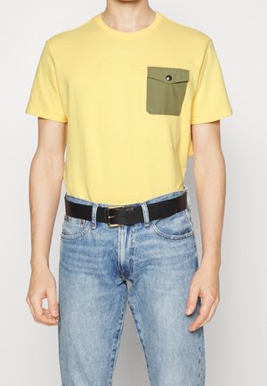 Homme portant un t-shirt jaune à manches courtes avec une poche poitrine verte boutonnée, un jean bleu clair et une ceinture en cuir noire avec une boucle dorée.