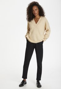 Pull beige en v avec une finition texturée, associé à un pantalon noir ajusté et des chaussures noires brillantes. Le pull présente une coupe décontractée.