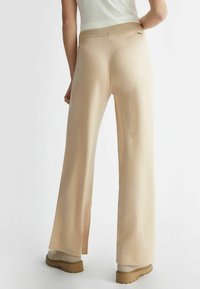 Pantaloni beige a gamba larga con una texture setosa e un cinturino elastico dorato; abbinati a calzature beige con suola testurizzata.