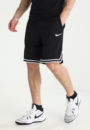 Homme portant un short Nike noir avec bordure blanche et des baskets Nike blanches avec des détails noirs, debout avec une main dans la poche.
