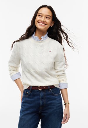 Young woman smiling wearing a white cable knit sweater over a blue striped shirt with dark blue jeans and a brown belt.