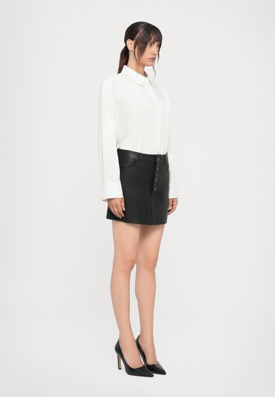 Chemise blanche à boutons associée à une mini-jupe en cuir noir, avec une fermeture à boutons à l'avant et des poches. Des escarpins noirs complètent le look.