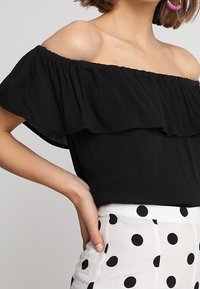 Haut noir épaule dénudée avec détail de volants superposés, en tissu léger, associé à un pantalon taille haute blanc à pois.