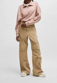 Jasnoróżowa bluza z kapturem i zamkiem, z białym nadrukiem, połączona z beżowymi spodniami cargo z kieszeniami i luźnym krojem, dopełniona białymi trampkami.